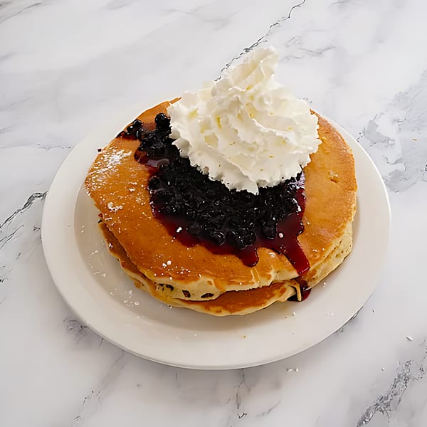 Fluffy pancakes topped with blueberry compote, whipped cream, and powdered sugar on a white plate.
